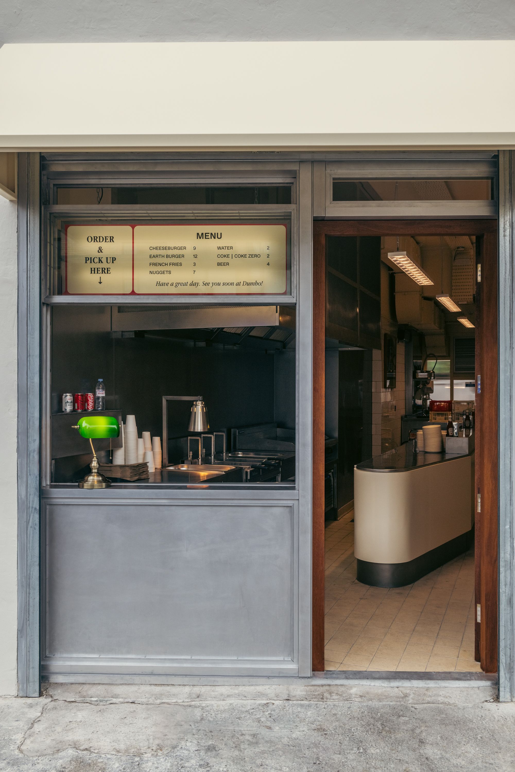 The exterior of a diner-style burger restaurant features a grey wooden frame, vintage back-lit banner showcasing products, a banker&#039;s lamp placed atop a counter, and buttery interiors mixed with chrome accents.