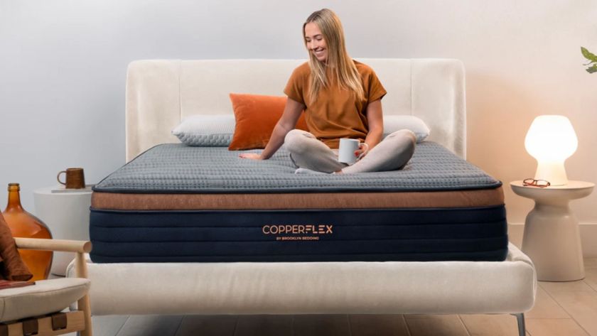 Blonde woman sitting crossed leg on Brooklyn Bedding CopperFlex mattress with mug in hand in bedroom with wooden floor and cream bed base