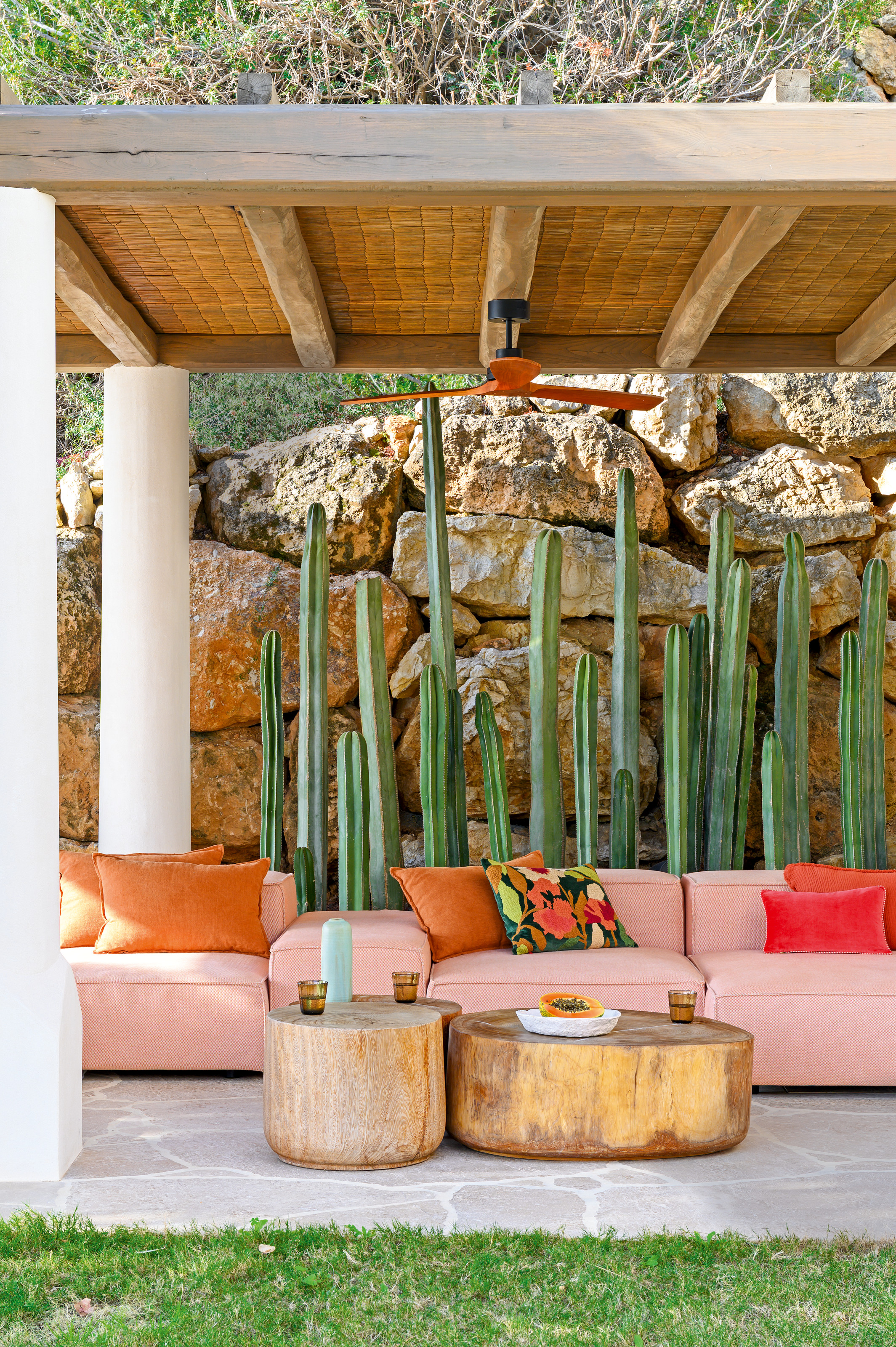 Outdoor lounge with pink modular sofa, wood dual coffee tables, cacti against a stone wall, all under a wooden pergola