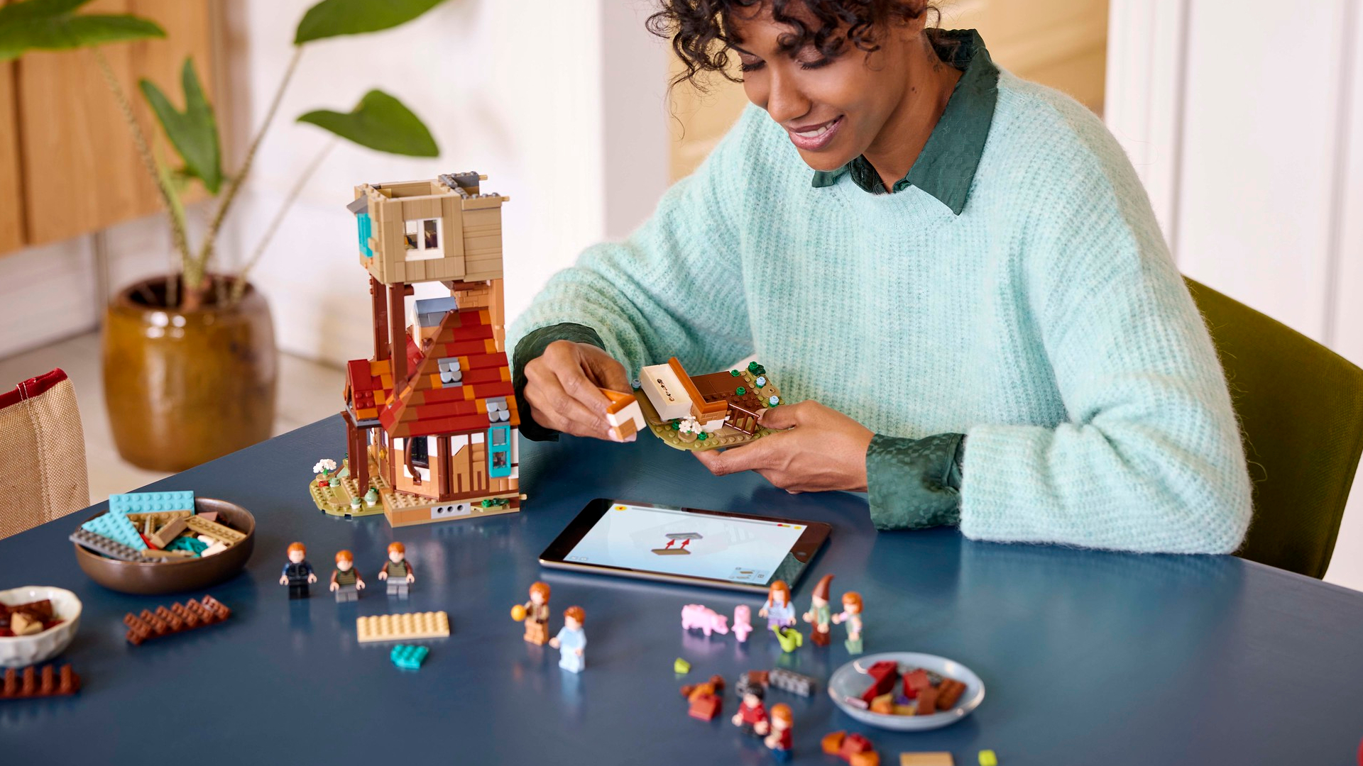 Lego The Burrow under construction by a woman sitting at a table, with bricks and minifigures arrayed around her