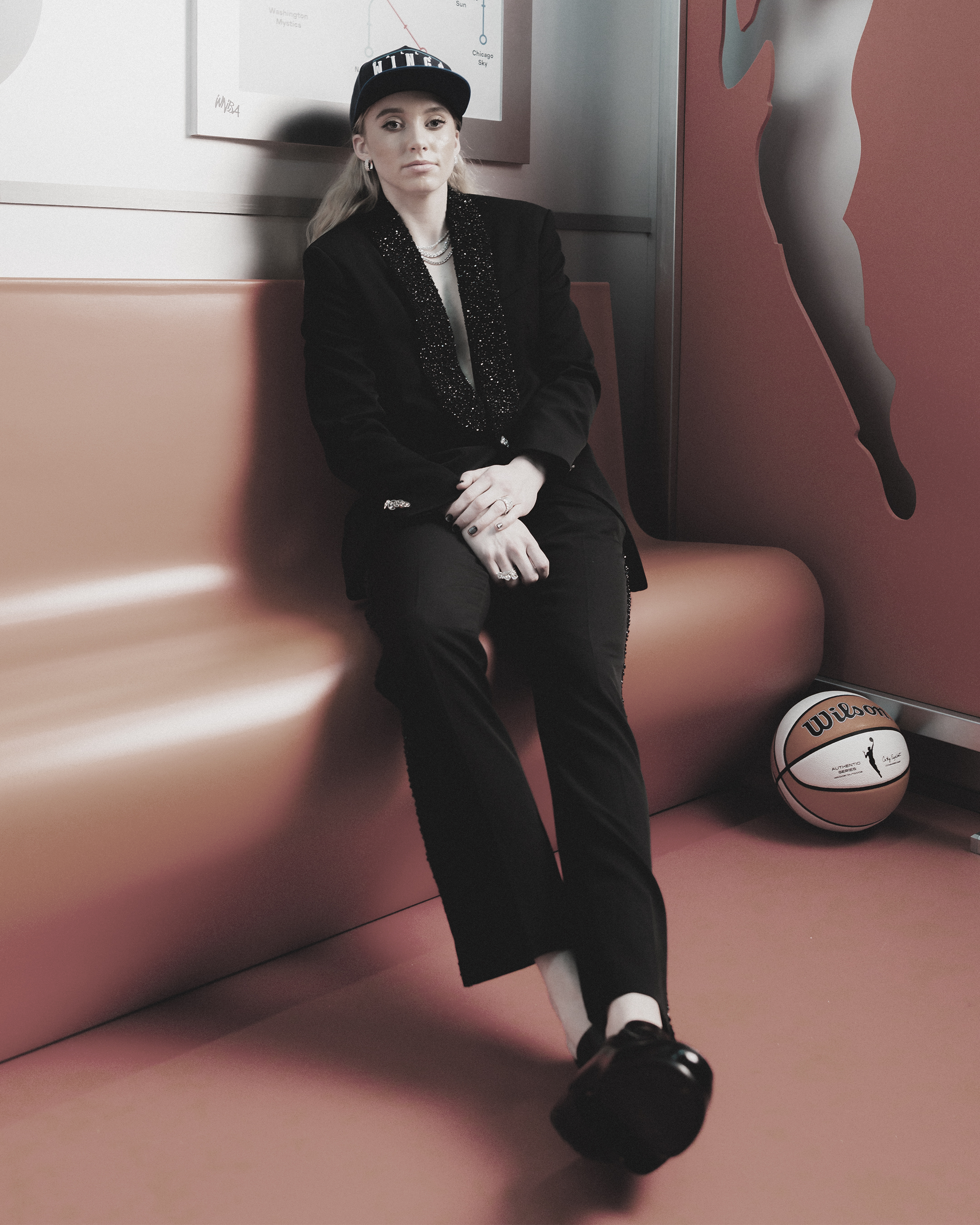 Paige Bueckers sitting on a bench in a black suit and cap, with a basketball at her feet.