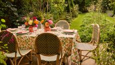 An outdoor dining table with a floral table cloth, two vases of flowers, and dinner plates laid out