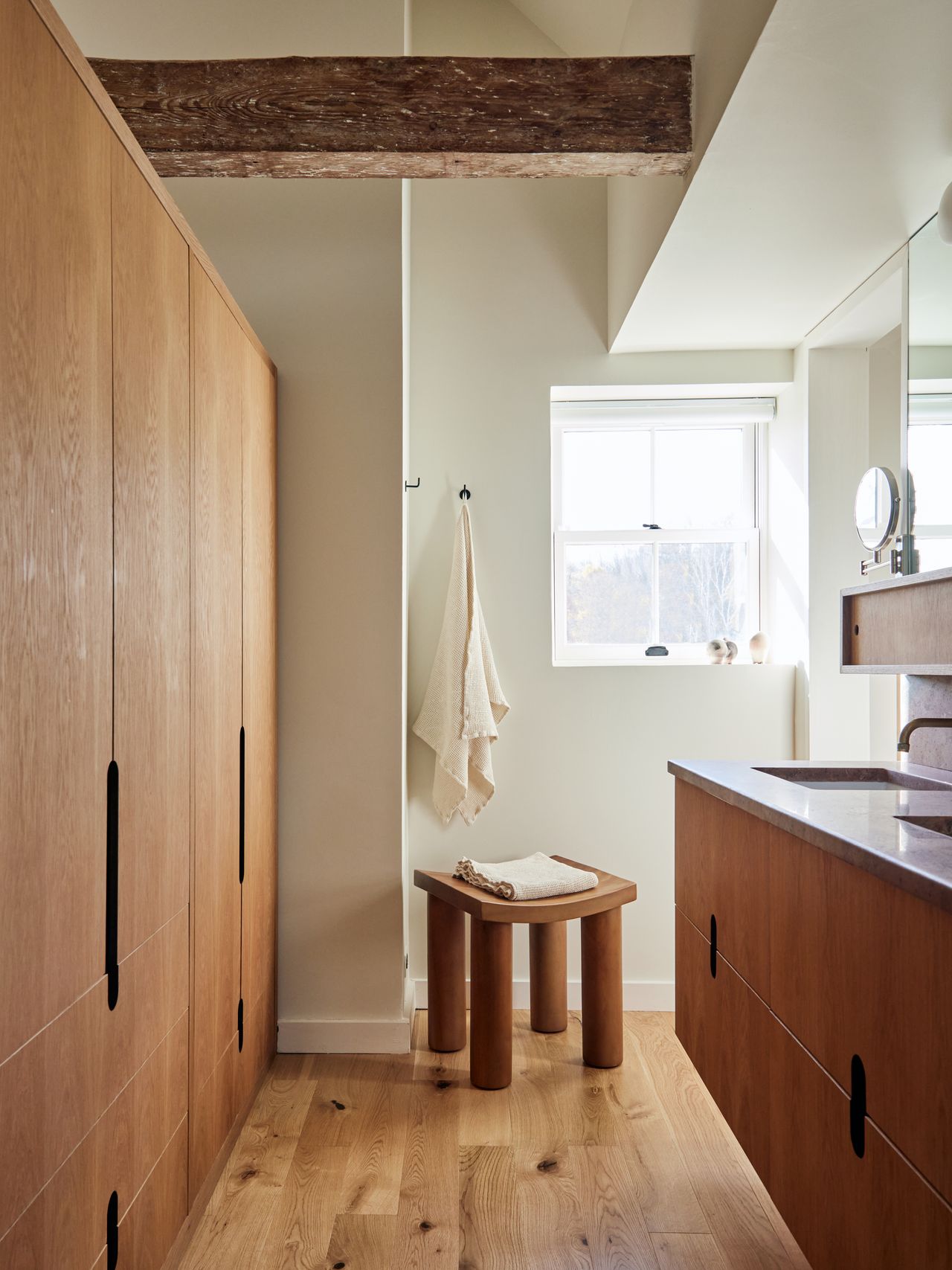 modenr bathroom with timber floor, timber cabinetry, floating timber vanity with stone top, two sinks, and mirror above. exposed timber beam in ceiling, and stool with folded towel