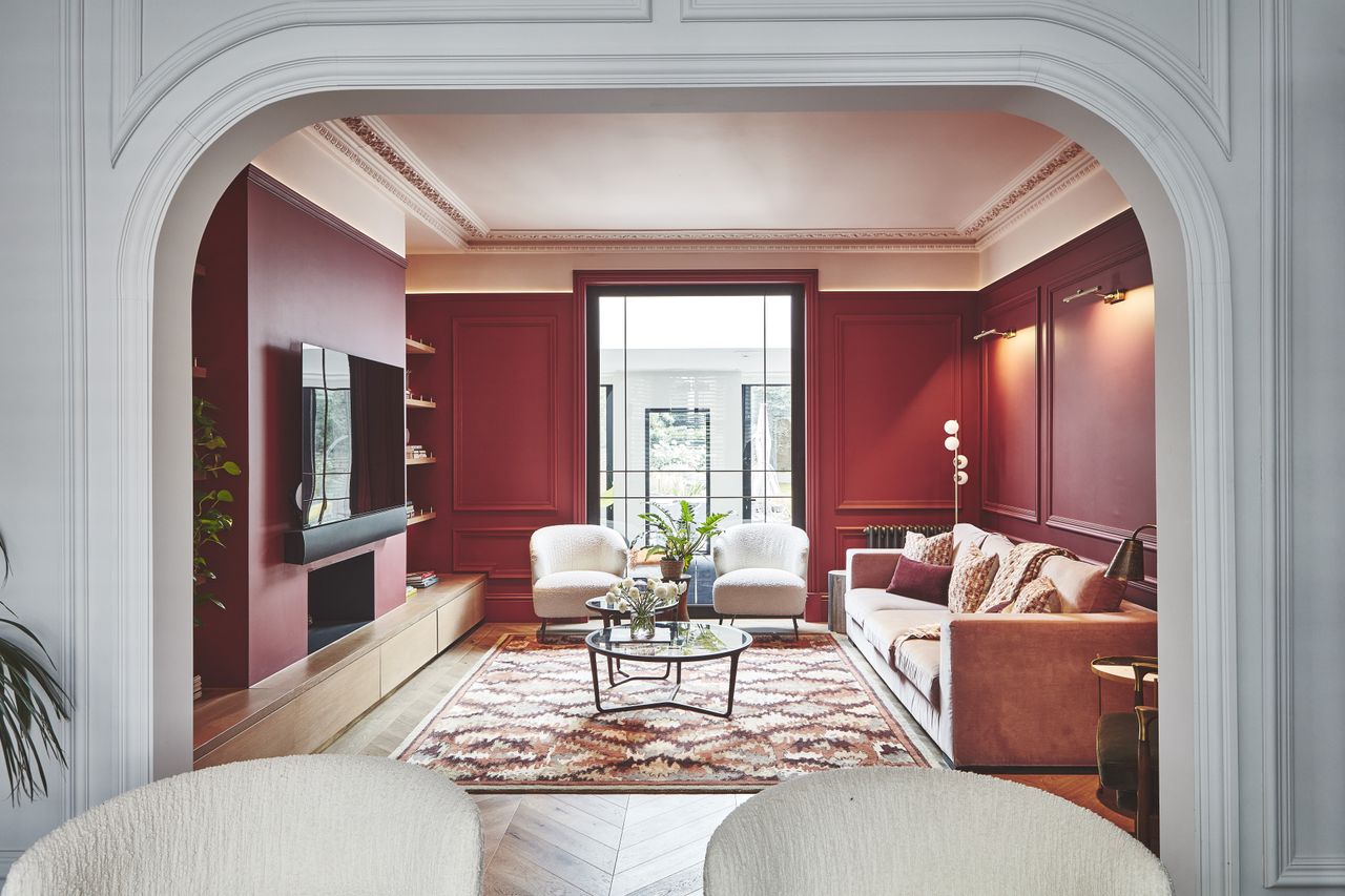 A view through a light blue architrave looking into a living room with dark red walls and earth toned furniture