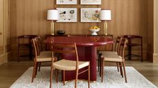 Image of a red lacquered dining table in a wood-drenched dining room. There are six wood and woven chairs around the table and a fluffy white rug on the ground.