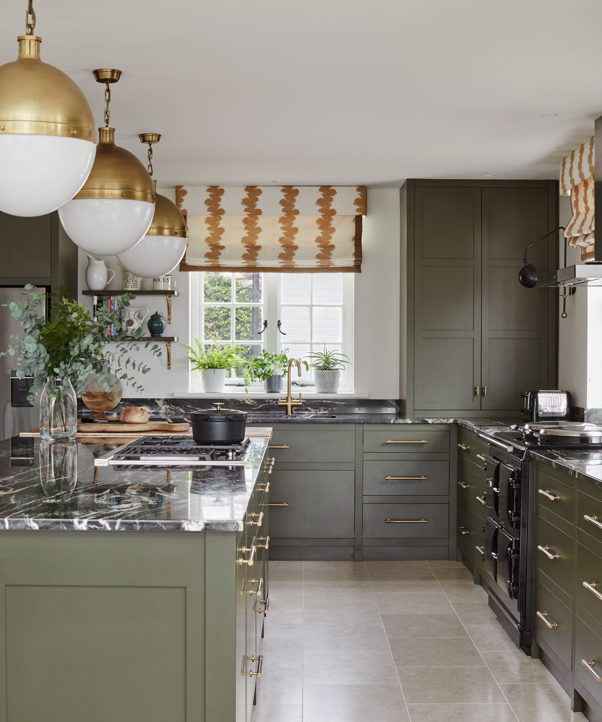 a green kitchen with dark marble countertops, gold cabinet pulls, three pendant lights, and striped blinds hanging in front of the windows