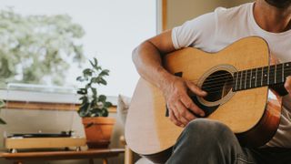 A man plays a chord on an acoustic guitar in a stylish, light home environment. Window behind provides a space for copy