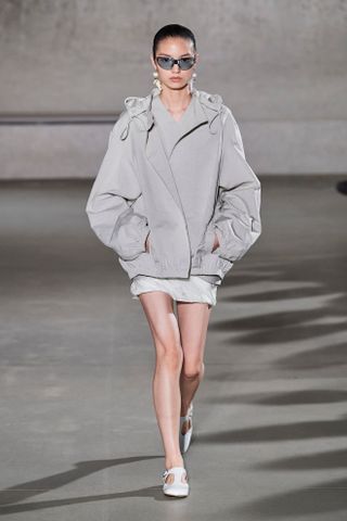 An SS24 runway photo at Tory Burch that shows a gray nylon sporty jacket with a gray mini skirt, blue sunglasses, and white T-strap flats.