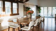 Farmhouse dining room ideas are beautiful. Here is a farmhouse dining room with dark wooden beams in a triangular shape at the top, a wooden dining table with white leather seats and a vase of roses, with large windows to the left