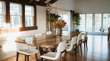 Farmhouse dining room ideas are beautiful. Here is a farmhouse dining room with dark wooden beams in a triangular shape at the top, a wooden dining table with white leather seats and a vase of roses, with large windows to the left