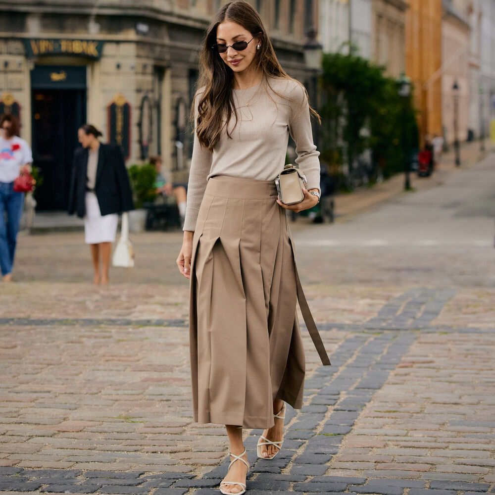 Woman wears pleated skirt, longsleeve top, and heeled sandals.
