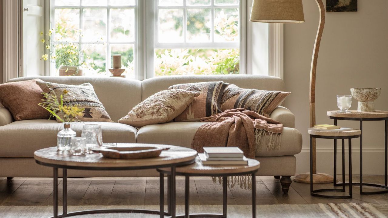 A spring themes living room with a large cream fabric sofa behind two round coffee tables. A glass vase of yellow flowers on the table. Assorted throw cushions on the sofa. A curved wooden floor lamp visible in the right hand corner. 