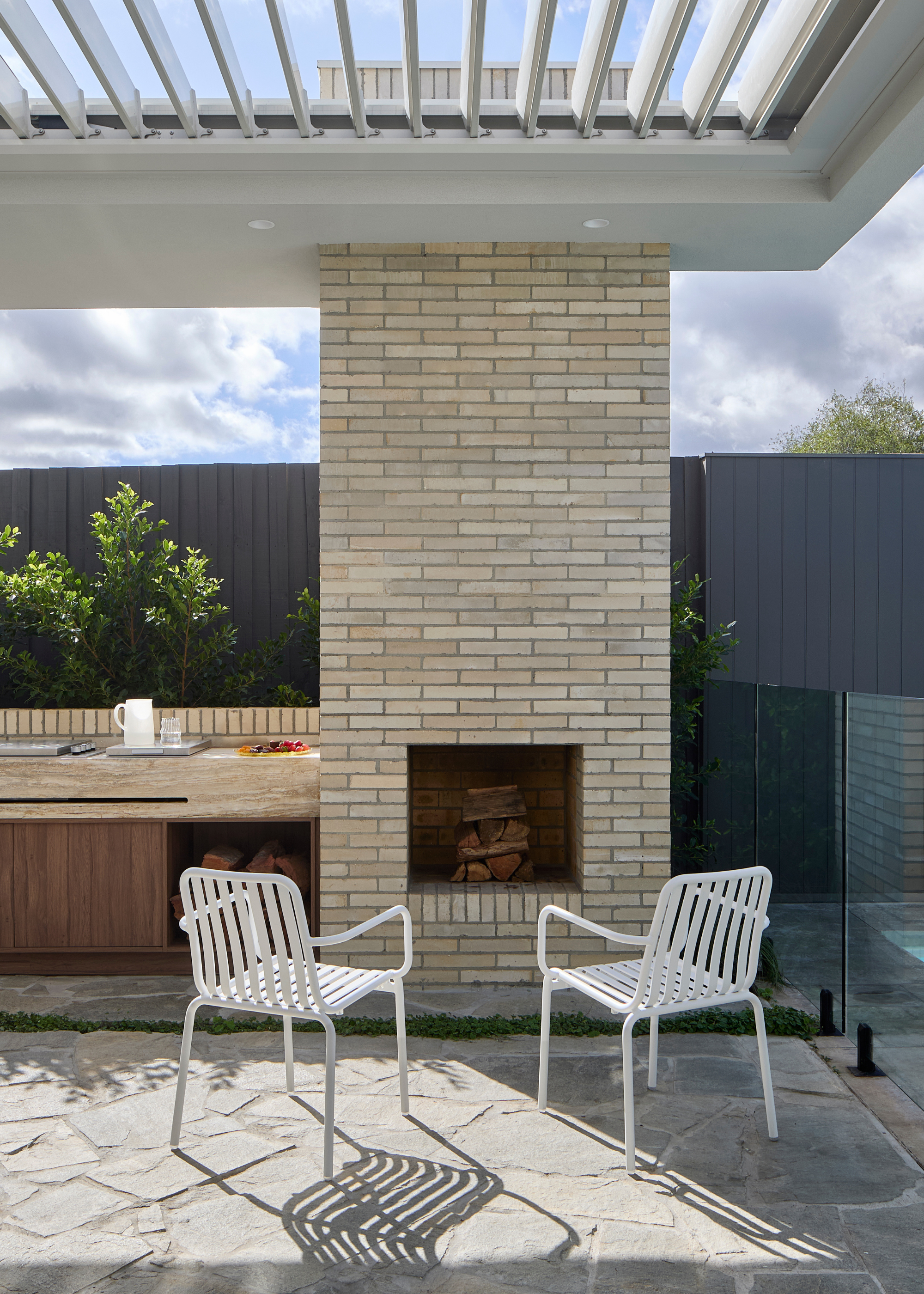 An outdoor kitchen with a built-in fireplace and a pair of white metal chairs