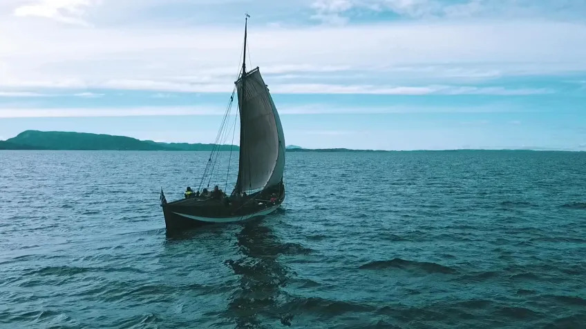 A small sailboat makes its away across turquoise waves towards the horizon, light blue with wispy white clouds