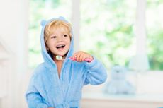 Little boy in blue bath robe or towel brushing his teeth in white bathroom with window on sunny morning