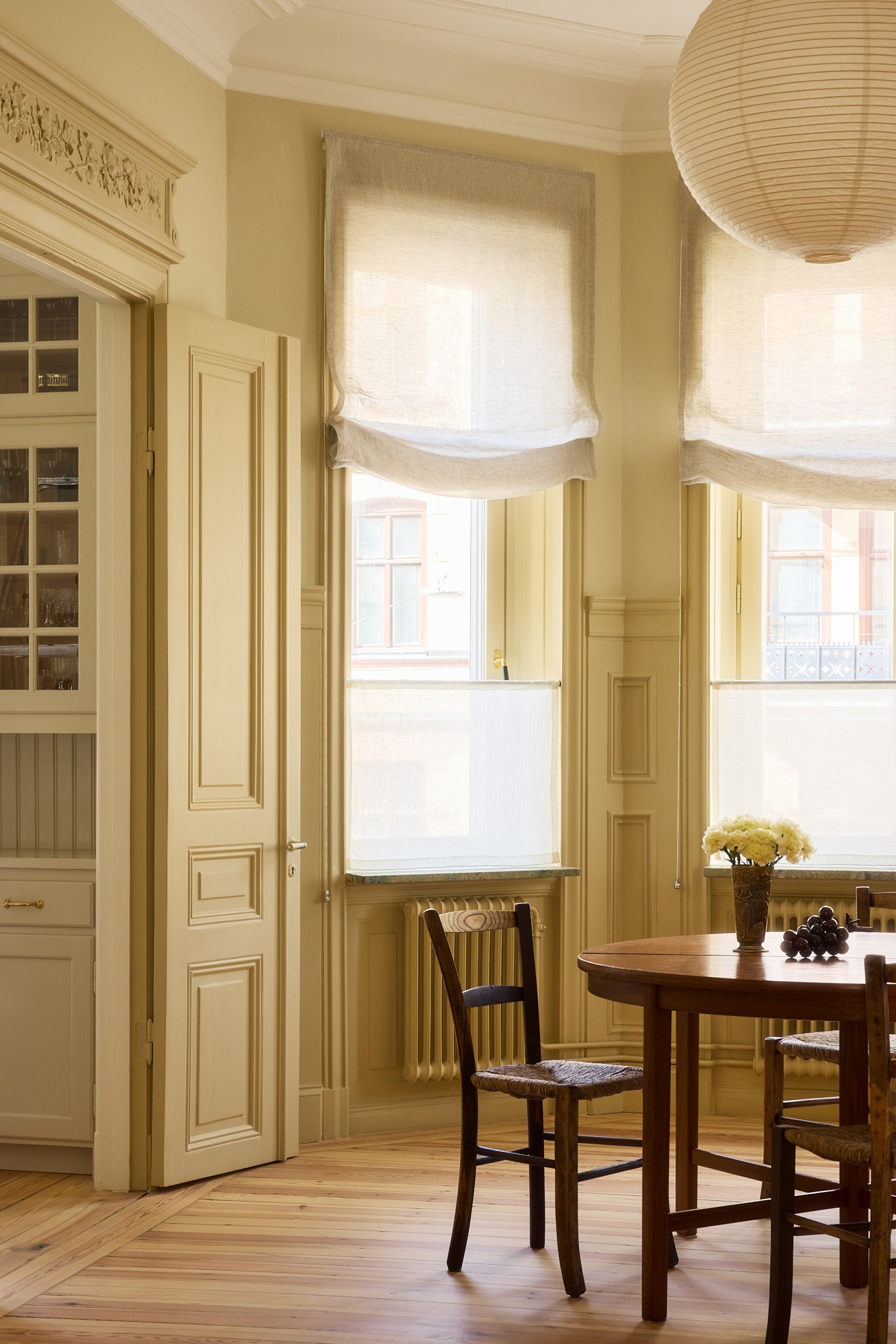 Soft Roman Blind Sheer Linen over bay windows in a beige dining space with crown molding on the walls. There is a wooden dining table and chairs with a paper lantern overhead.
