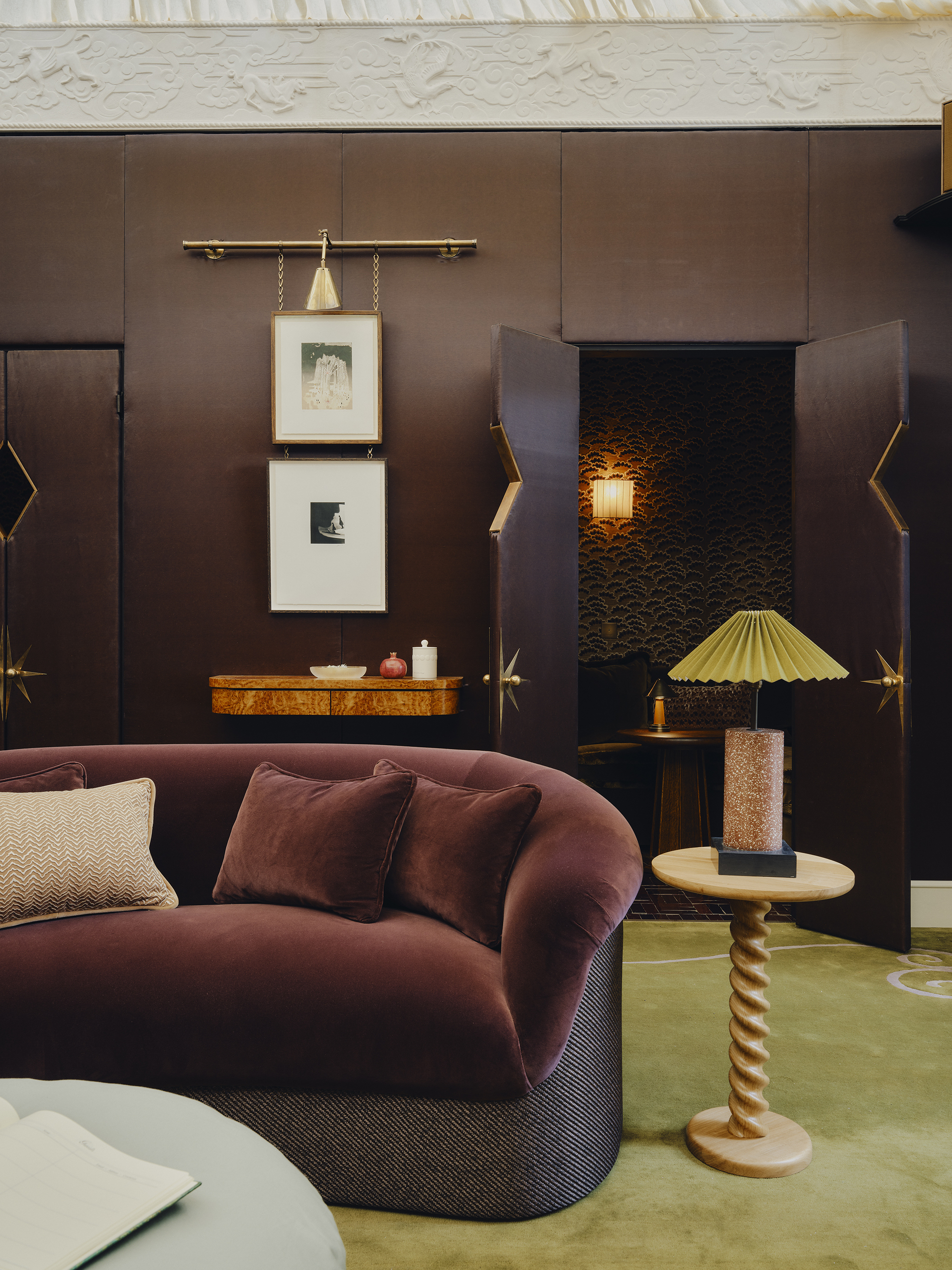 living room with chocolate walls, ornate cornice, bright green rug, velvet brown curved sofa, artwork hung on picture rail, timber side table, and doors leading through to a bar