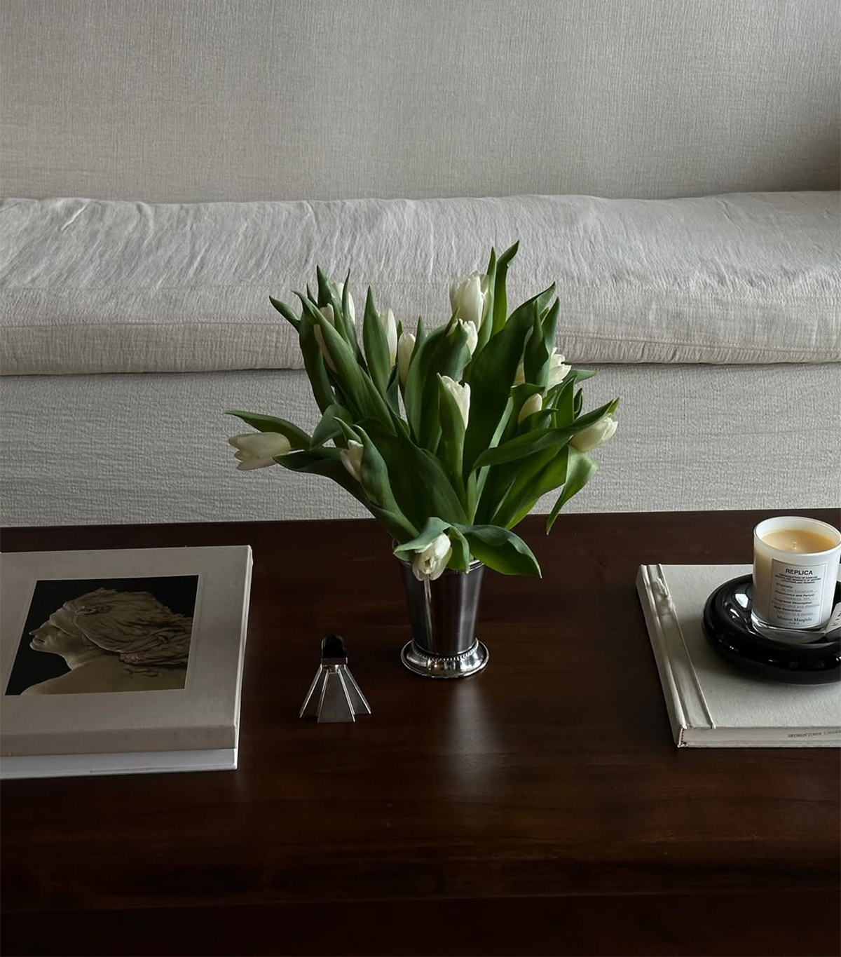Dawn Tan&#039;s wood coffee table and white linen couch with flowers, a coffee table book, and a candle.