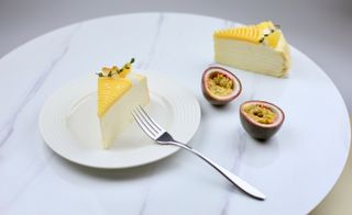 Pancake cake slices are displayed on a white marble table. One slice is set directly on the table, while the other is on a white plate. A passion fruit sliced in half sits in between.