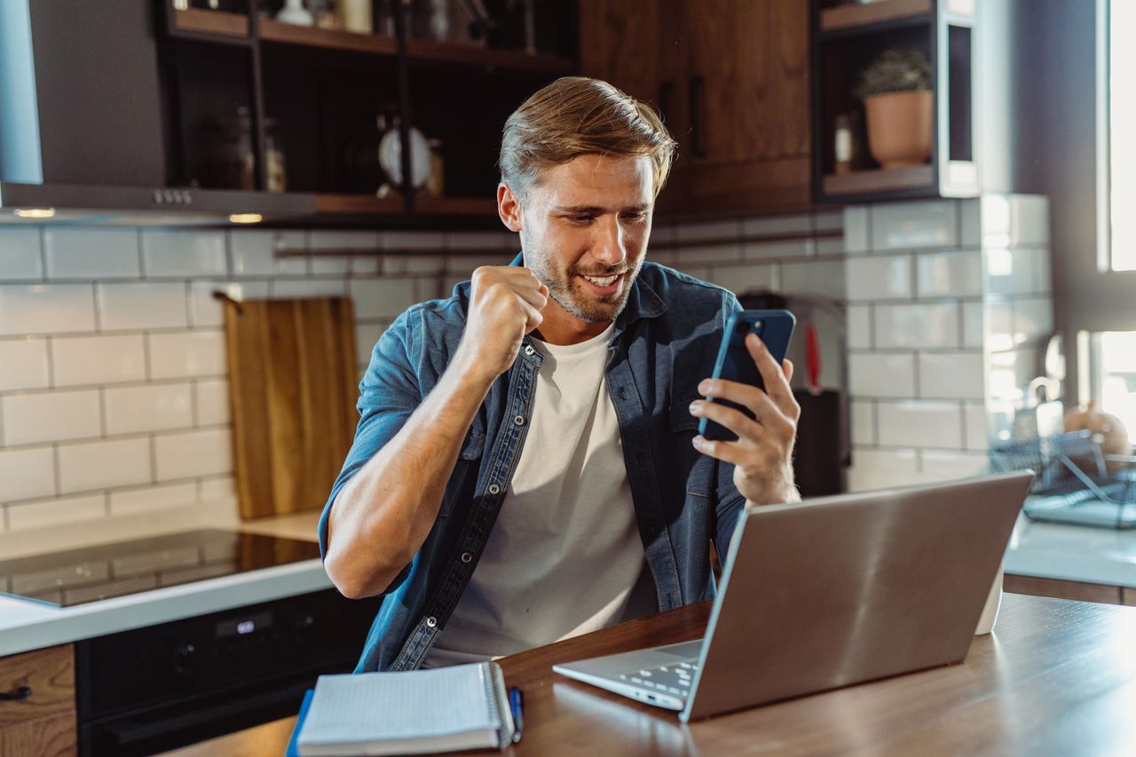 A smiling man is sitting in the kitchen with a laptop and mobile phone while receiving good news