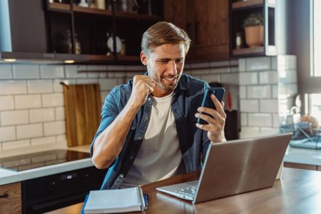 A smiling man is sitting in the kitchen with a laptop and mobile phone while receiving good news