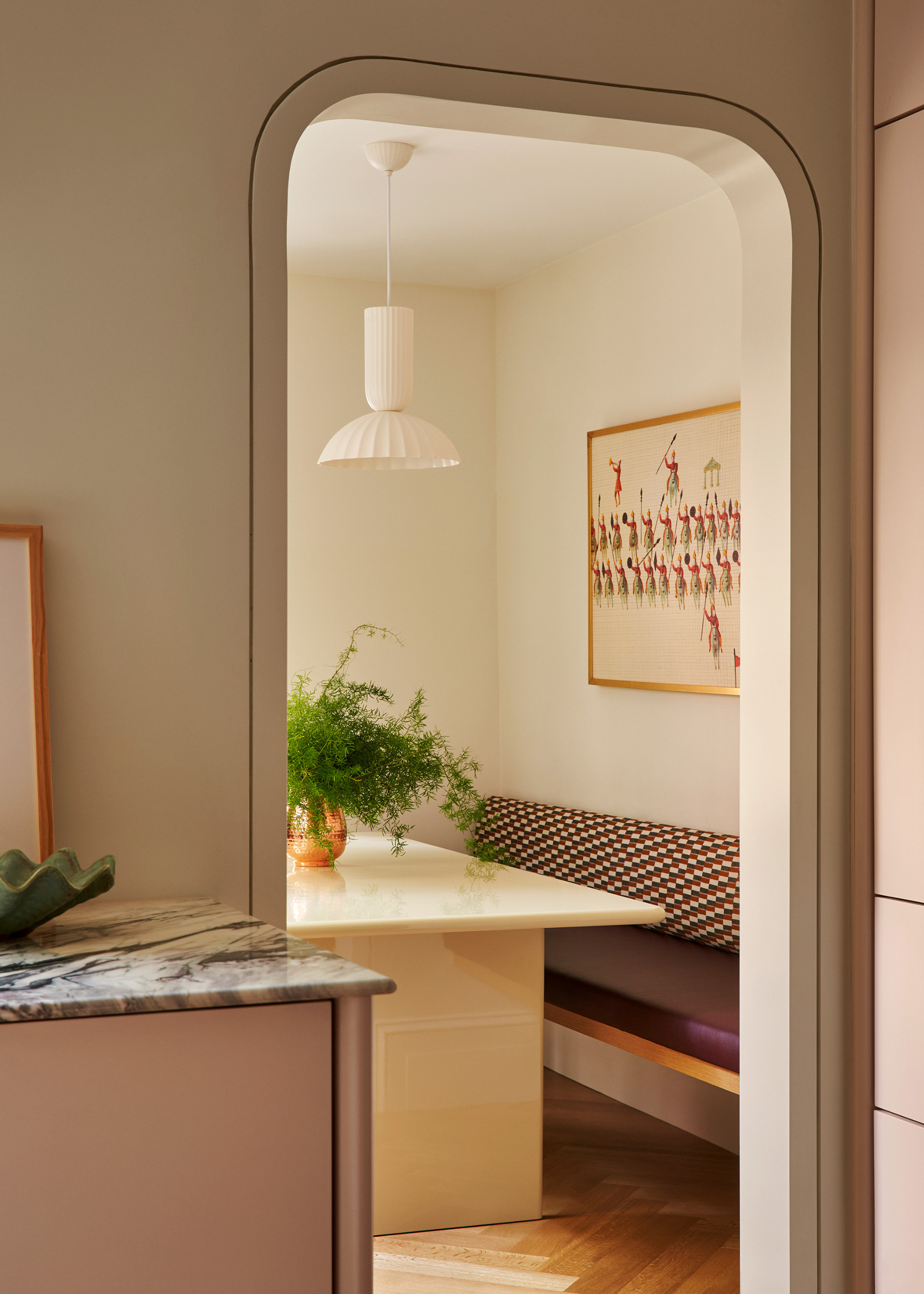 A kitchen with an arched doorway leading to a dining nook with a stone table and a vase of stems, a white pendant lamp above, a burgundy and patterned seat, and a hanging framed painting