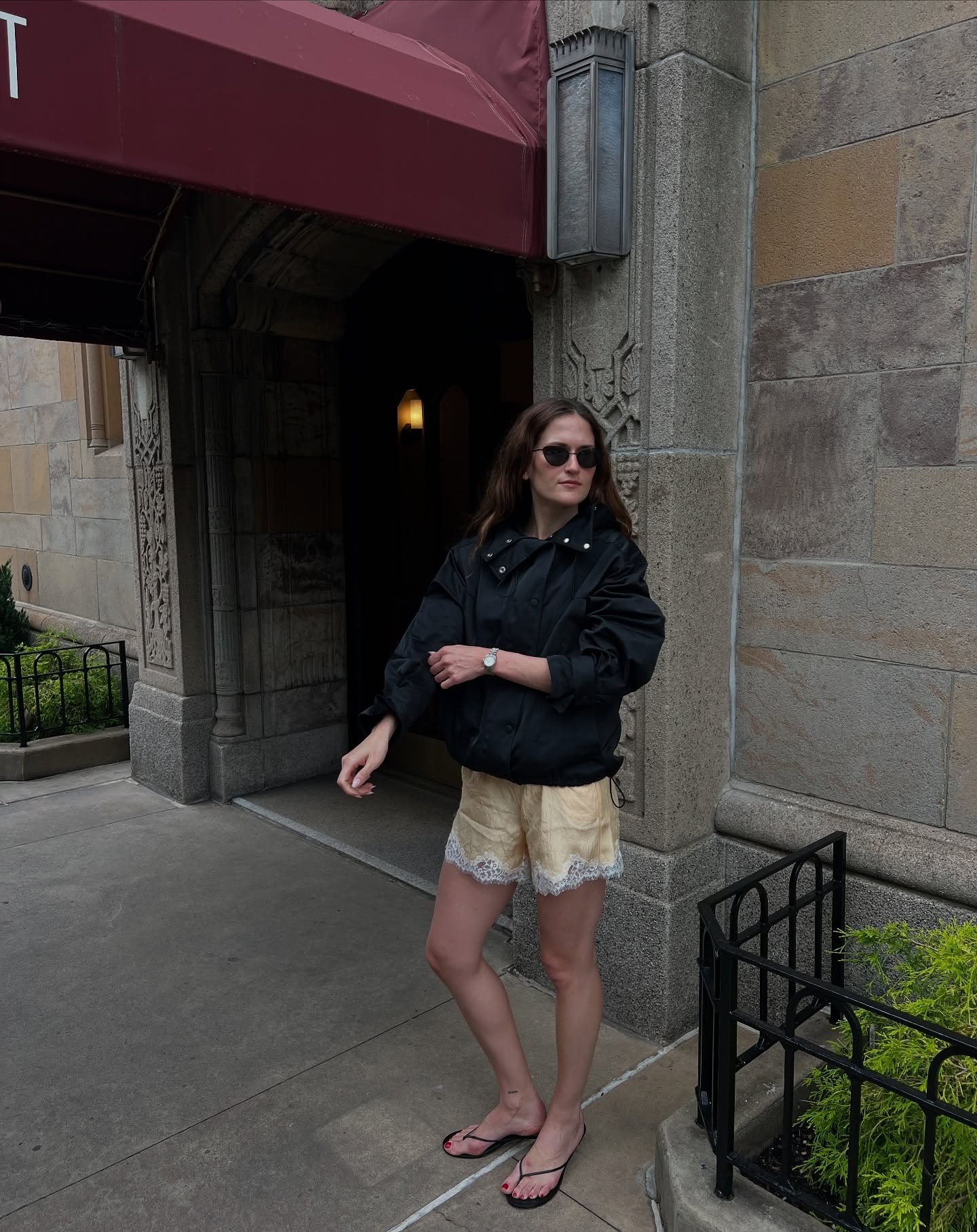 Woman wearing black windbreaker with lace yellow shorts, black flip flops, standing on New York City street.