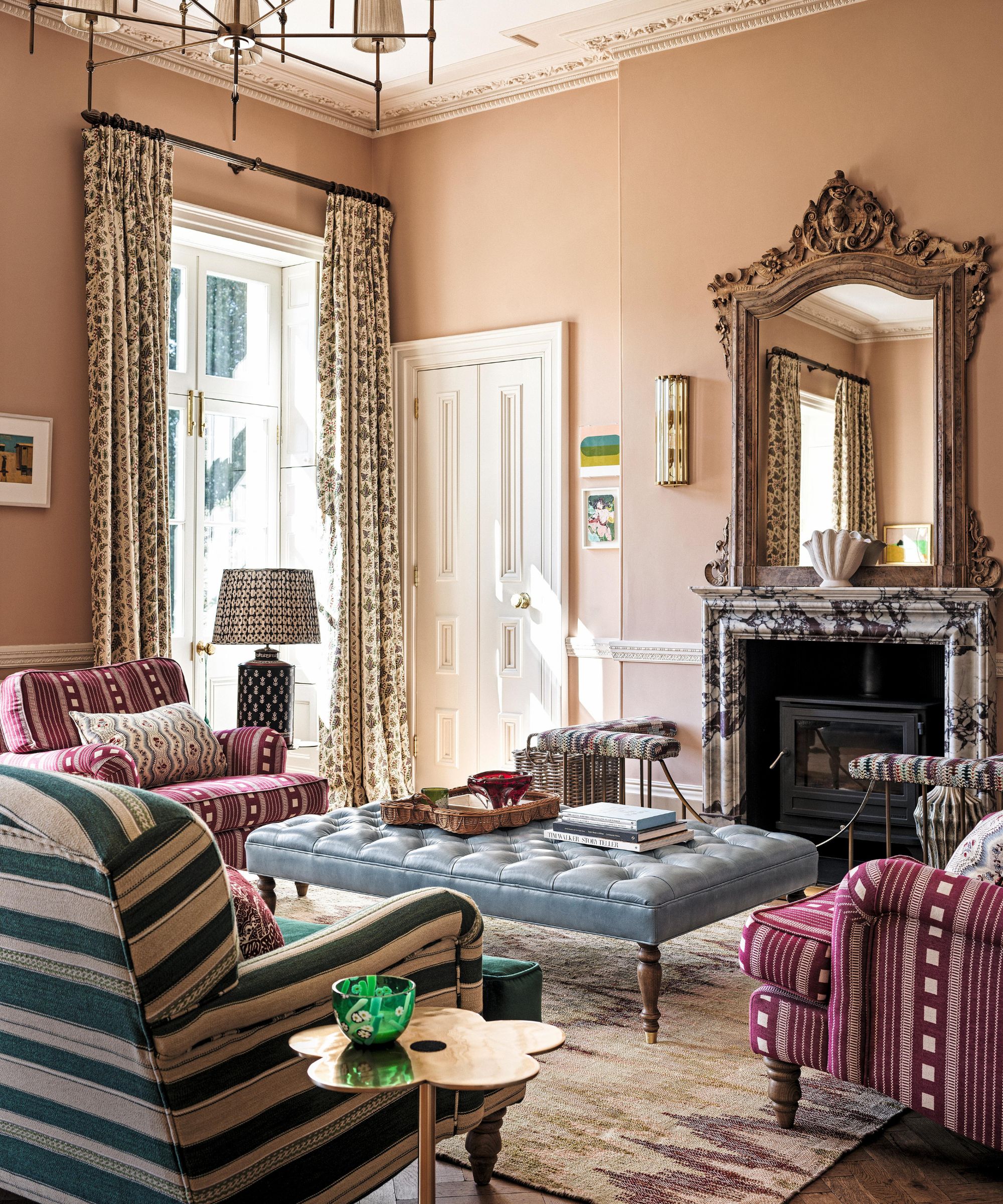 a warm pink traditional english sitting room with marble fireplace, ornate mirror, large windows, striped armchairs and a blue ottoman