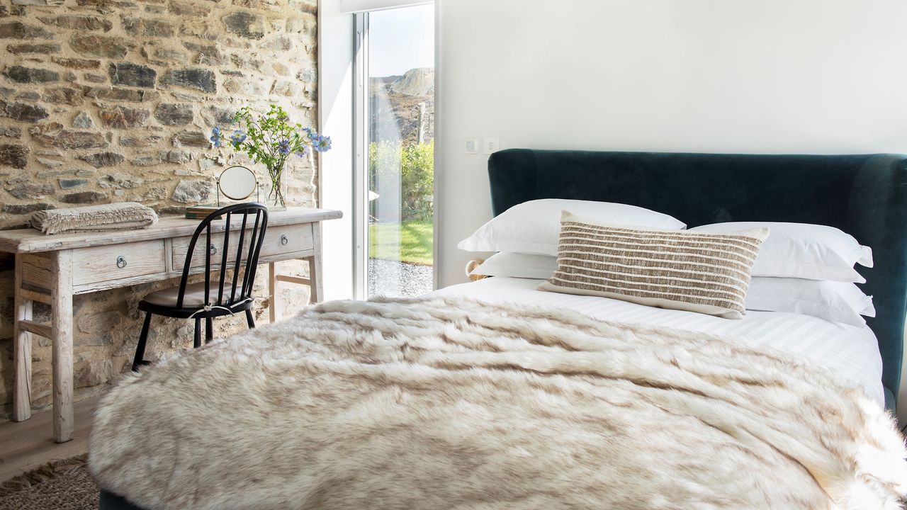 White duvet set with furry throw blanket on bed, white rustic desk and chair with stone feature wall in white painted bedroom