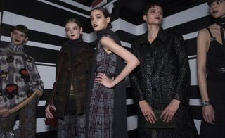 Five female models wearing looks from Bottega Veneta's collection. One model is wearing a patterened neck scarf, grey jumper with multicolour design, grey and brown patterned trousers and she is holding a bag. Another model is wearing a dark coloured high neck jumper, brown plaid style jacket, grey patterned trousers and she has a dark red bag on her shoulder. The third model is wearing a grey patterened piece and dark grey scarf. The fourth model is wearing a black sparkle effect coat and she is holding a dark green clutch bag. And the fifth model is wearing a black patterned dress and black neck scarf