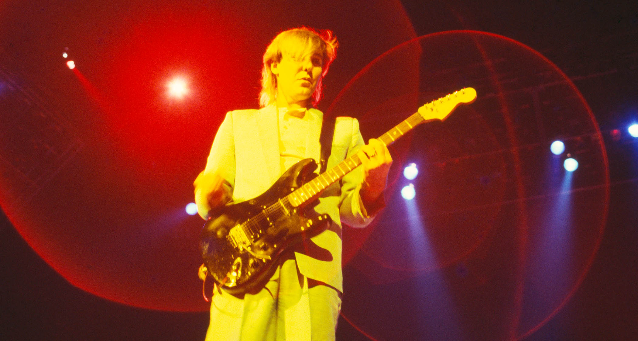 Alex Lifeson of Rush plays his all-black Hentor Sportscaster onstage in 1985. He wears a grey suit.