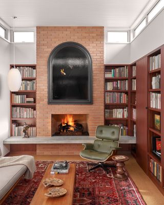 A mid-century style living room with red-toned wood bookshelves, brick fireplace, red Persian rug and green mid-century armchair