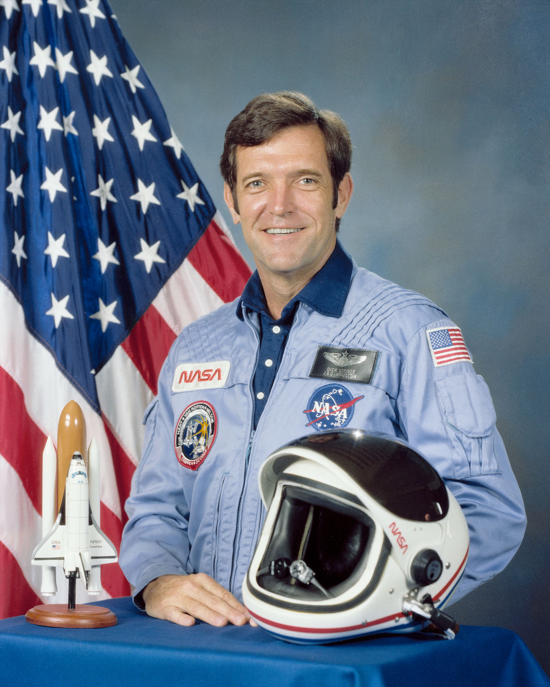 a man in a blue flight suit stands in front of an american flag