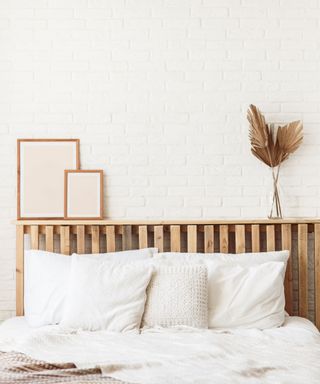 A light brown wooden headboard with white sheets and pillows in front of it and two framed pink wall art prints and a slim vase of brown dried flowers above it, and a white brick wall behind it