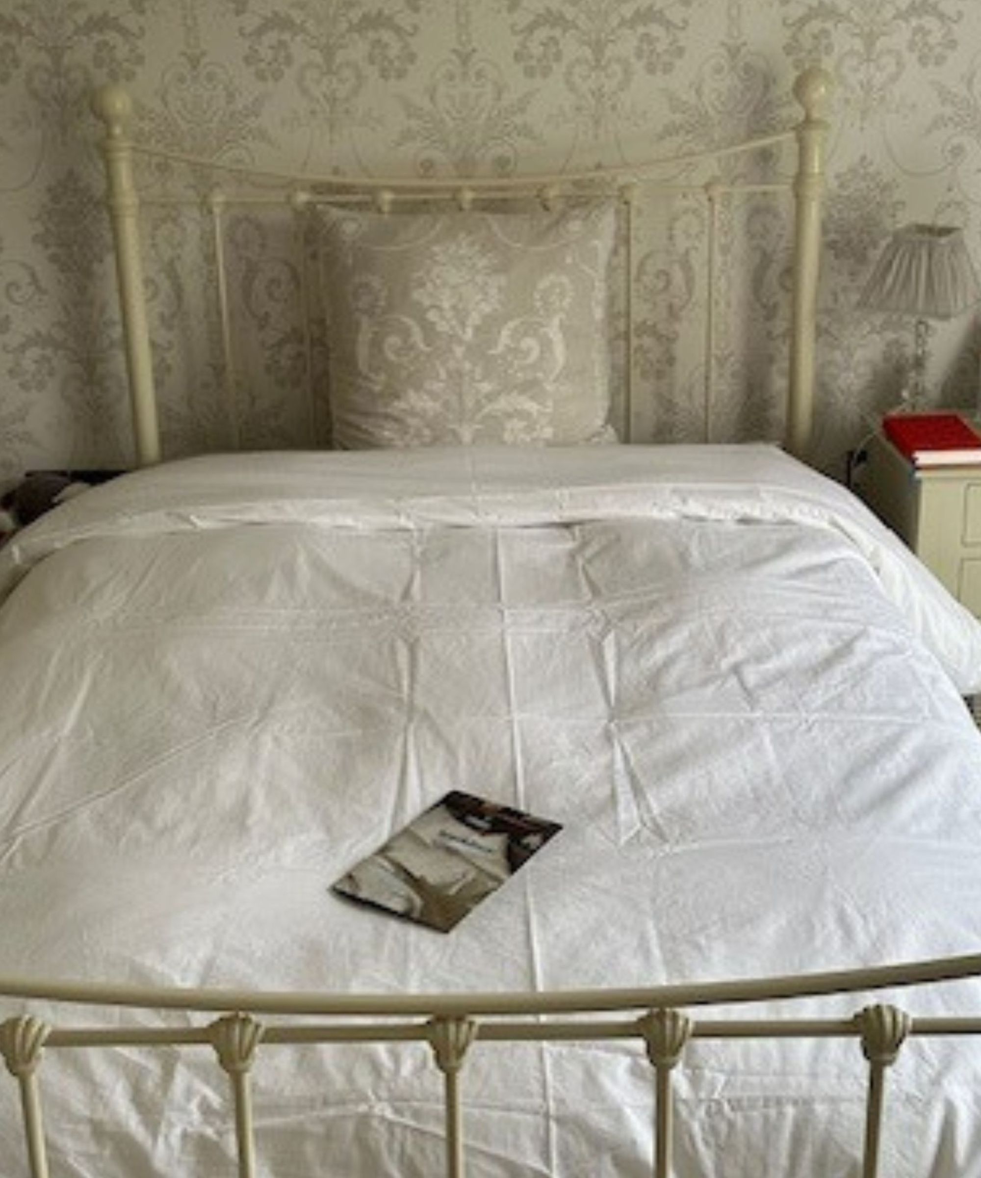 The Brooklinen down duvet in a white cover on a white framed bed, with a silver cushion and a book in the center of the bed. The wallpaper behind is cream with silver patterning, and there's a bedside table to the right with a red book, and small plastic lamp with grey lampshade.
