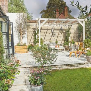 Patio area with a pergola and a seating area underneath it, surrounded by grass and plants