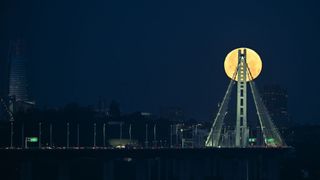 a bright full moon shines behind a tall triangle shape supporting bridge structure.