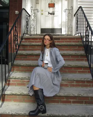 A woman wears a gray blazer, white t-shirt, gray pleated skirt, and black engineer boots.