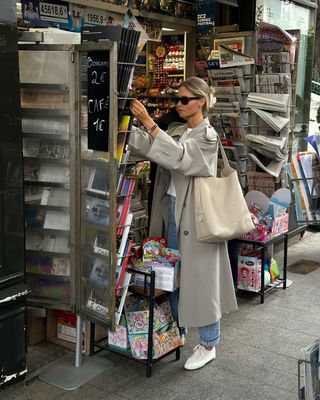 @clairerose carries a cream shoulder bag whilst wearing a grey trench coat, blue jeans and white trainers