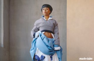 Myha’la poses wearing a grey cropped sweater and blue floral skirt against a grey and beige wall, with marie claire logo in bottom right corner for Marie Claire September 2024 issue