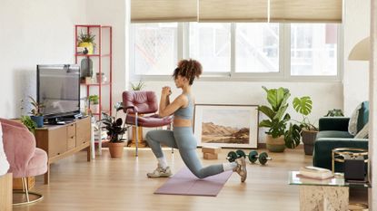 Woman doing lunges while watching online yoga class. Female is wearing sports clothing in living room. She is working out at home.