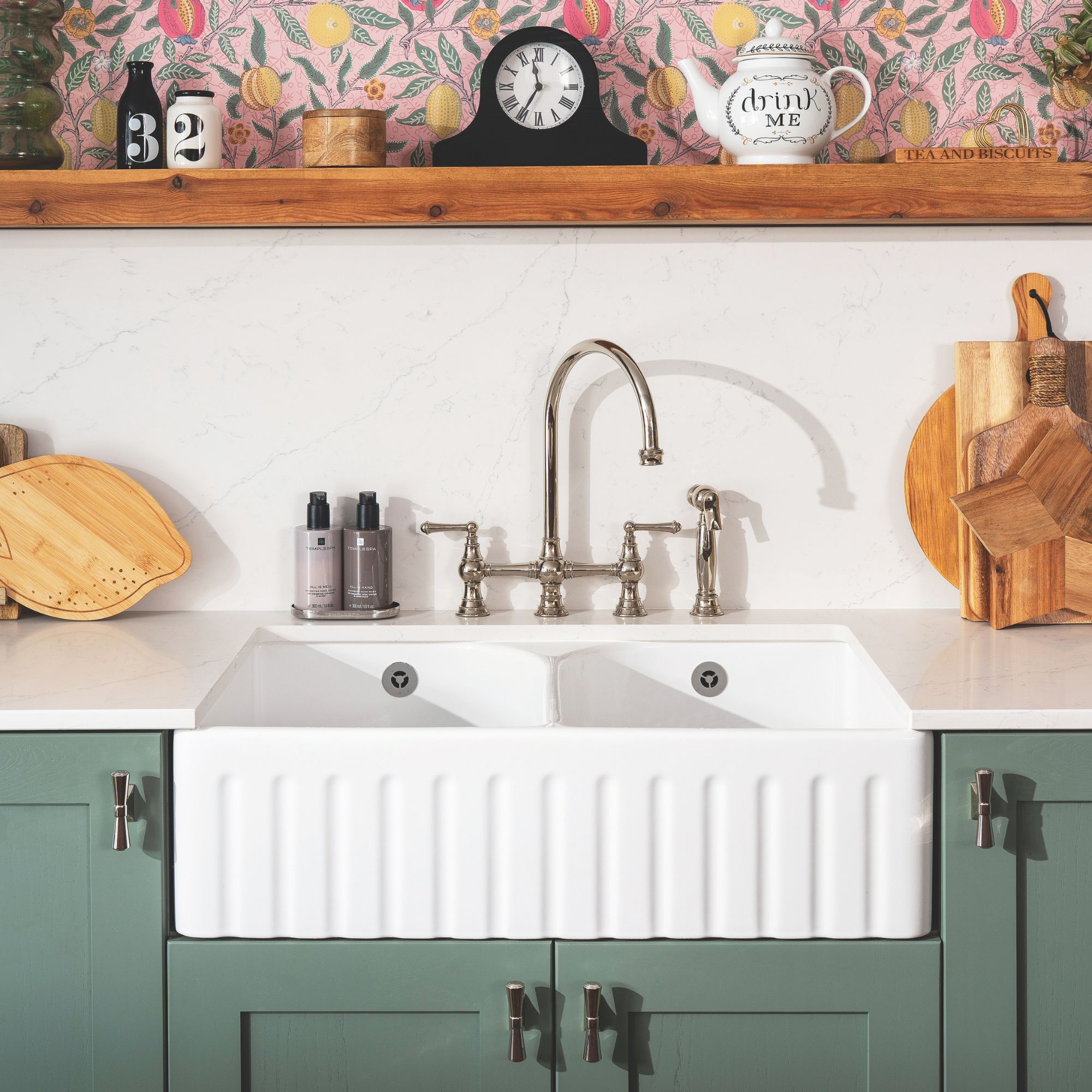 White painted kitchen with green cabinets and a white ceramic sink, with bright floral wallpaper above the splashback