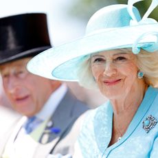 Queen Camilla in a turquoise hat and coat sitting next to King Charles