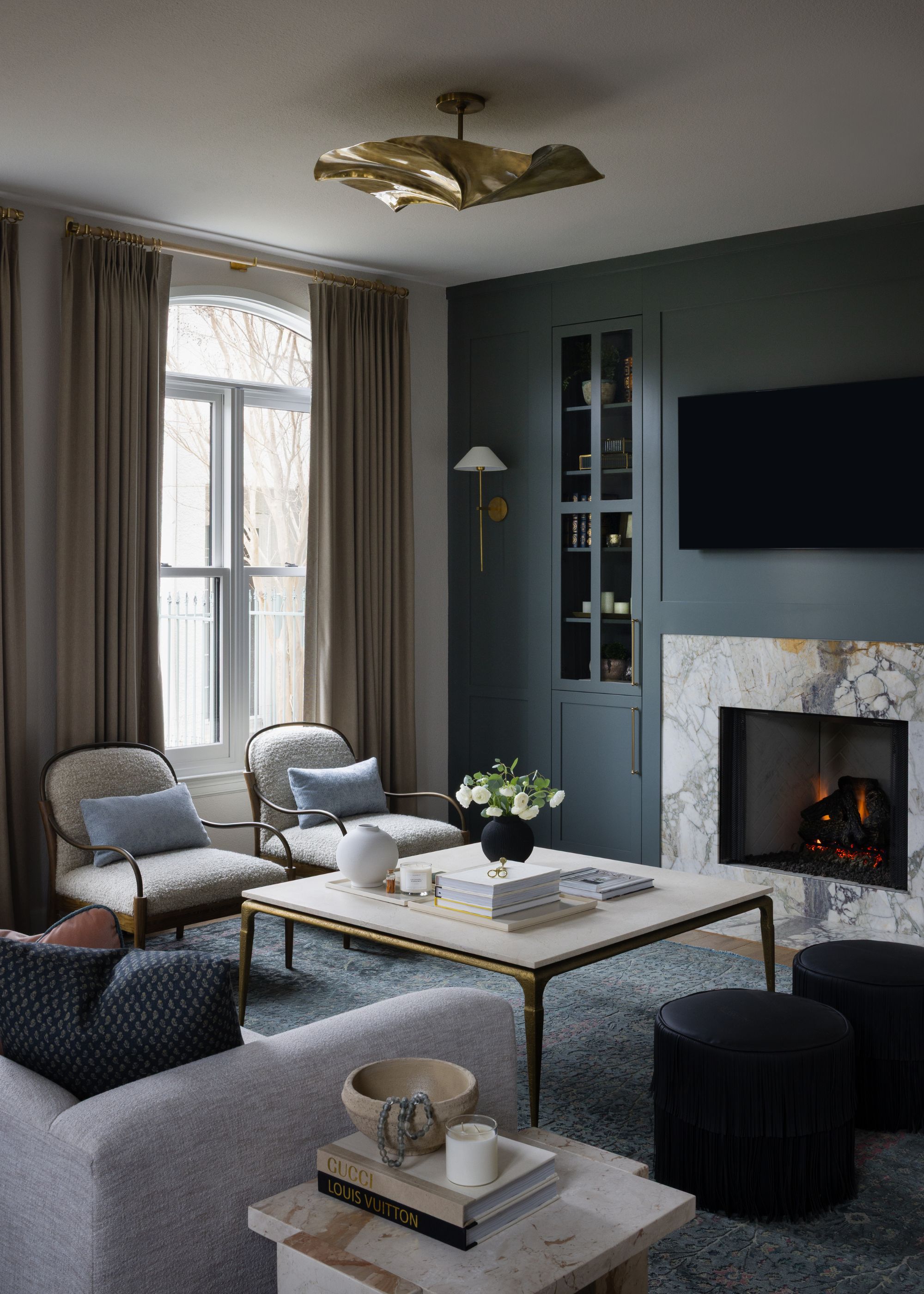 A moody, sophisticated living room featuring a dark teal built-in bookcase and a marble fireplace with a crackling fire. Two cream boucl&amp;eacute; armchairs with blue pillows face a large stone coffee table.