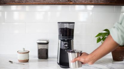 OXO Conical Burr Coffee Grinder on a white countertop with a hand inserting the grounds container underneath. Tehre are coffee beans at the side and a sugar pot too