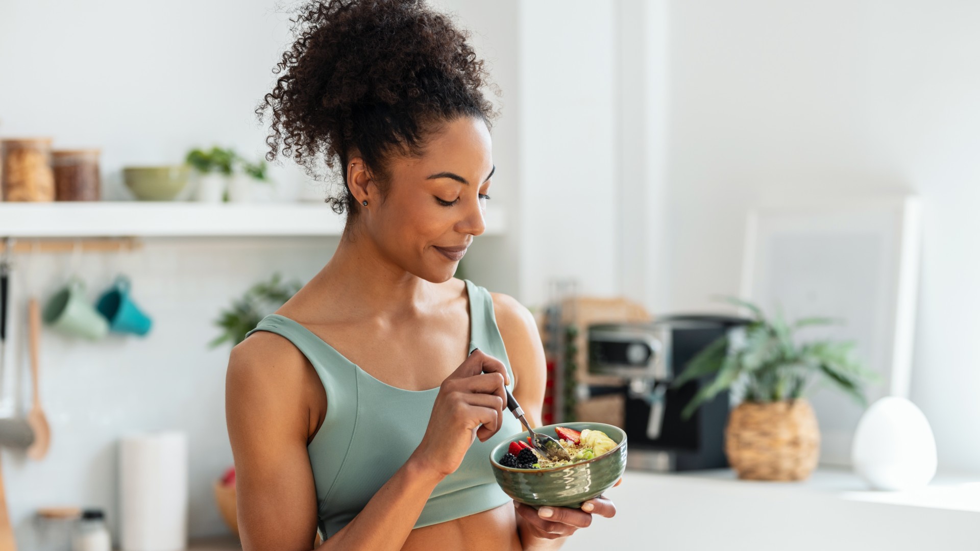 Woman eating a bowl of food in a light and airy kitchen wearing a sports bra
