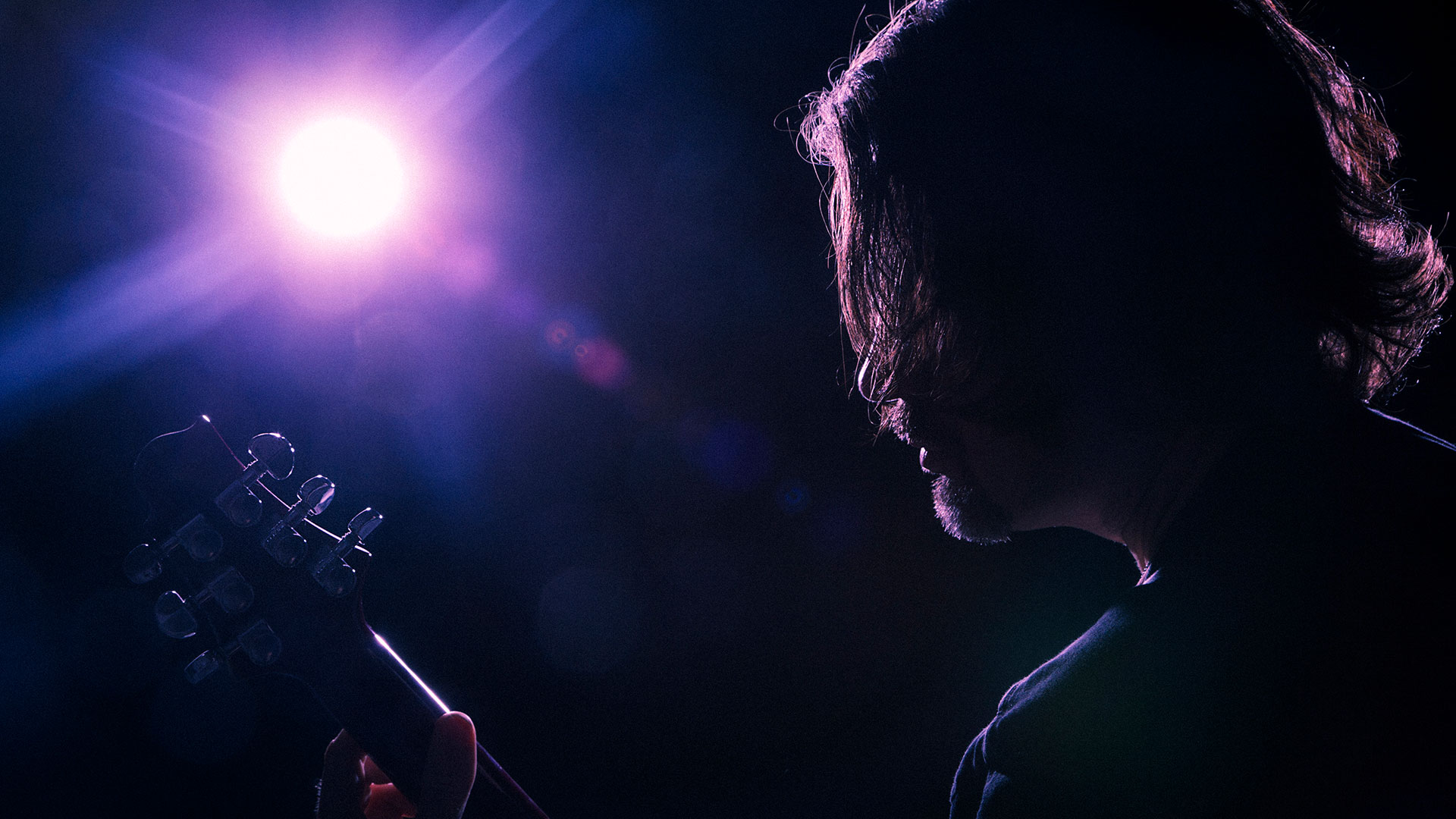 A silhouetted guitar player with a stage light in the background. Photographed directly into the light to create flare