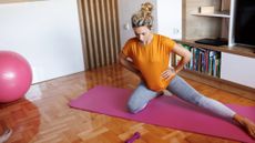 Woman kneels on one knee on exercise mat, with her other leg extended to the side. She is in a domestic setting, with shelving and a TV behind her