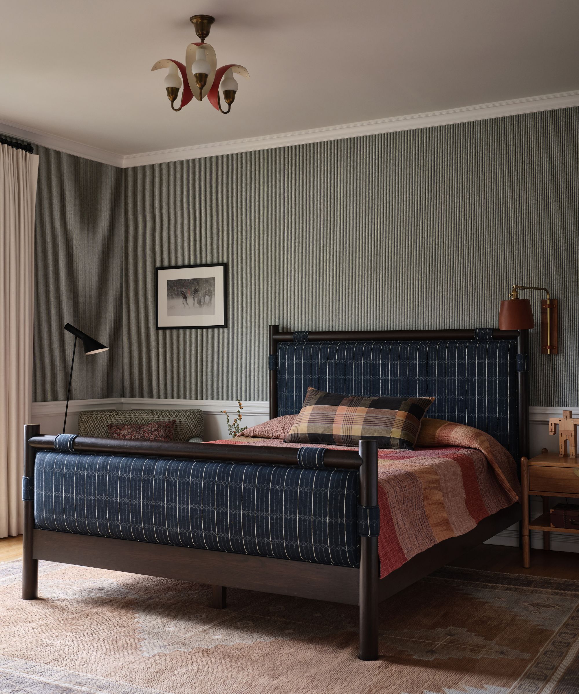 Bedroom with dark vertical striped wallpaper, white wainscoting, a dark wood and navy woven bed frame, and a brightly colored plaid throw blanket
