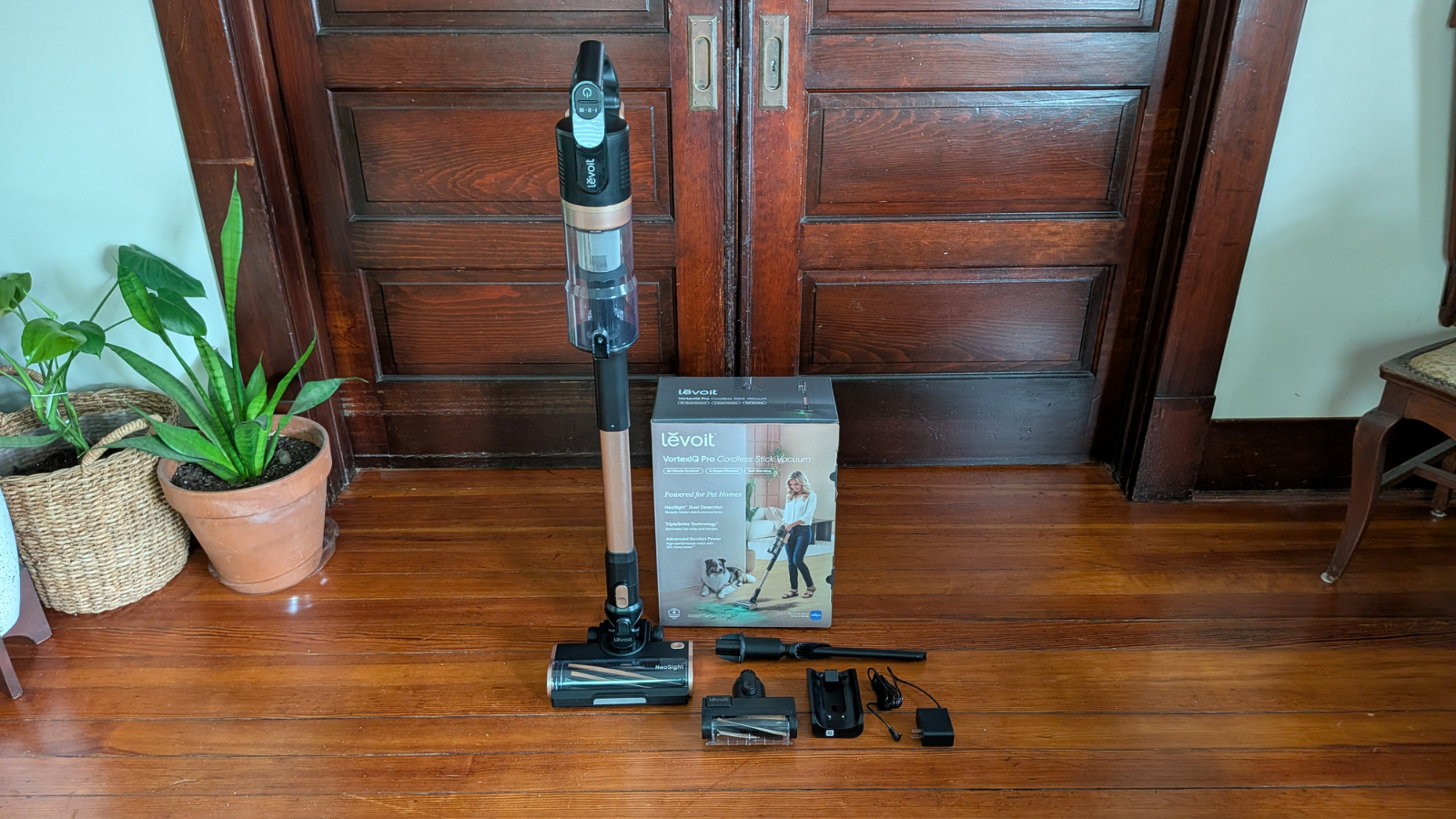 The Levoit VortexIQ Pro vacuum cleaner stands upright alongside the box it came in and the attachments and charging base. 